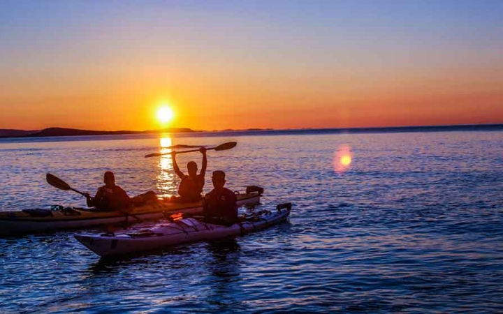 two kayaks rest in blue water while the sun sets behind them. one kayaker raises their paddle in the air in celebration.